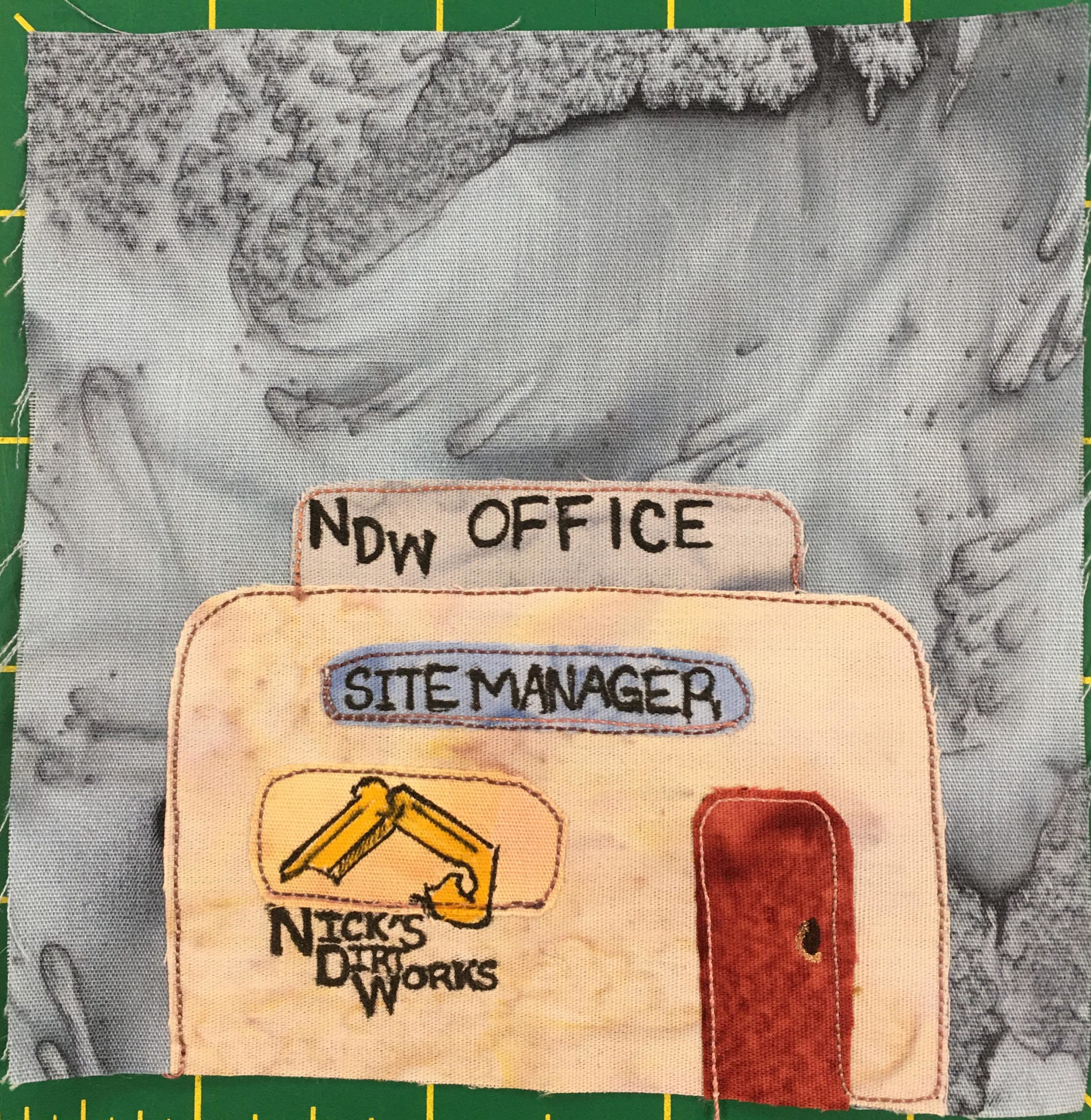 This quilt block shows a small office building against a light gray background. The building looks like a temporary construction office with a sign sticking out of the top of the roof that reads,