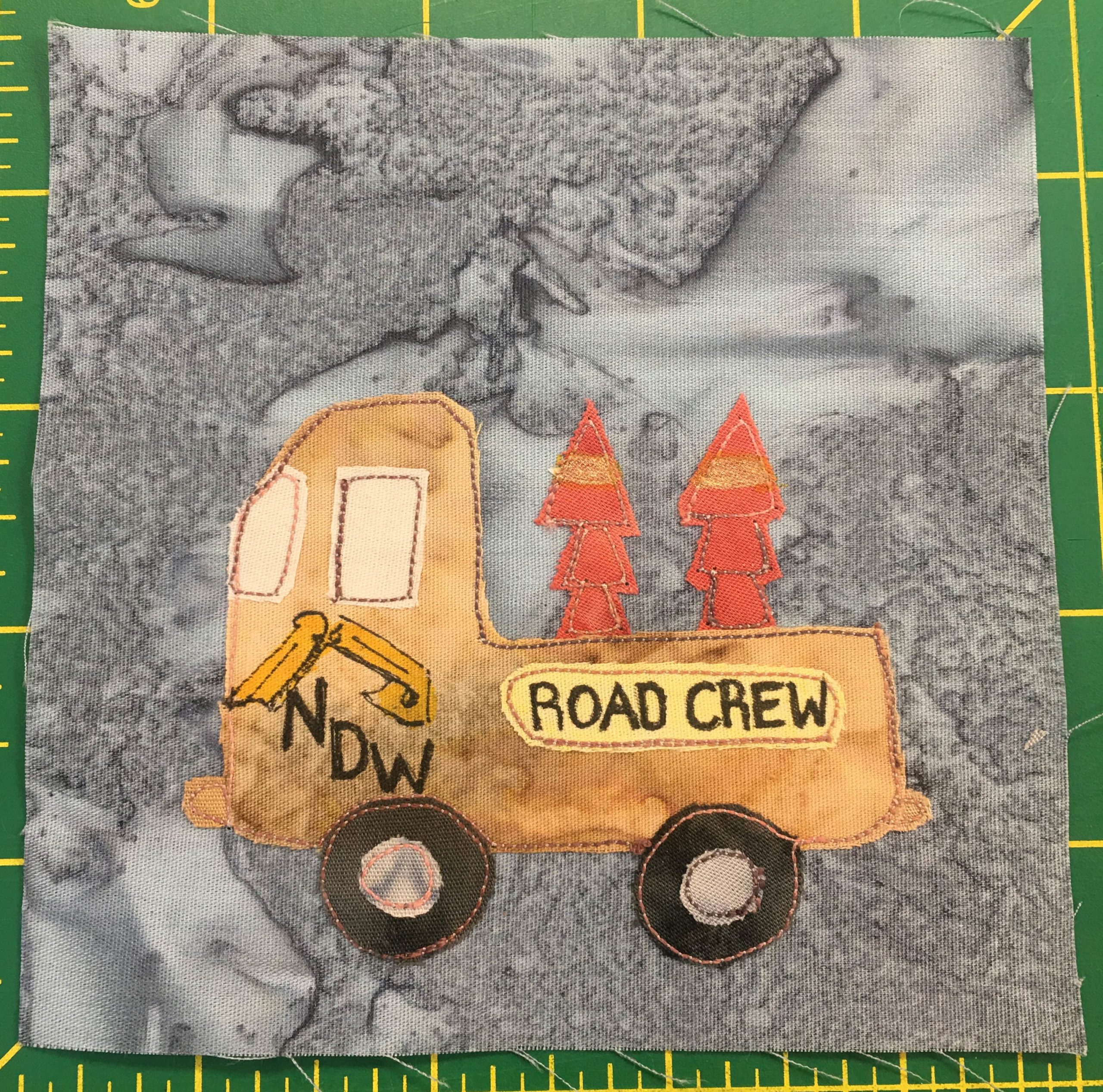This quilt block shows the side-view of a truck with a deep bed that contains stacked orange traffic cones. On the side of the truck are the words,