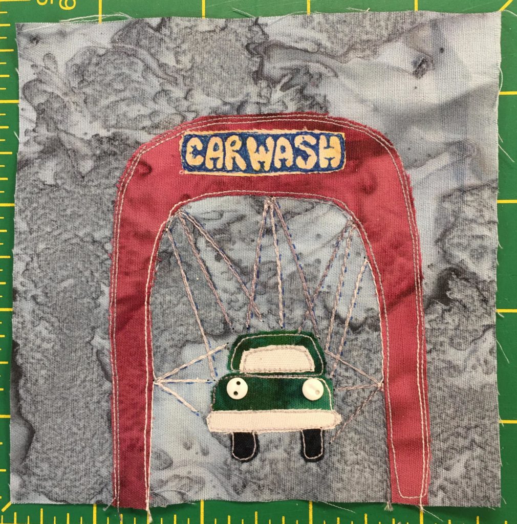 This quilt block shows a drive through carwash from the front. The canopy covering is in a maroon color and on the top is a sign in yellow on blue that reads,
