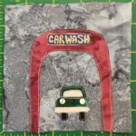 This quilt block shows a drive through carwash from the front. The canopy covering is in a maroon color and on the top is a sign in yellow on blue that reads,