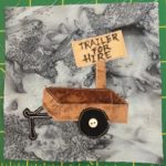 This quilt block shows a little box trailer that you might attach to a small car or truck against a gray background. The trailer is brown with a single wheel axis. The wheel is black with a small white button in the center to indicate it's attachment to the axis. There is a light brown wooden sign behind the trailer that looks like it's been written on by hand that reads,