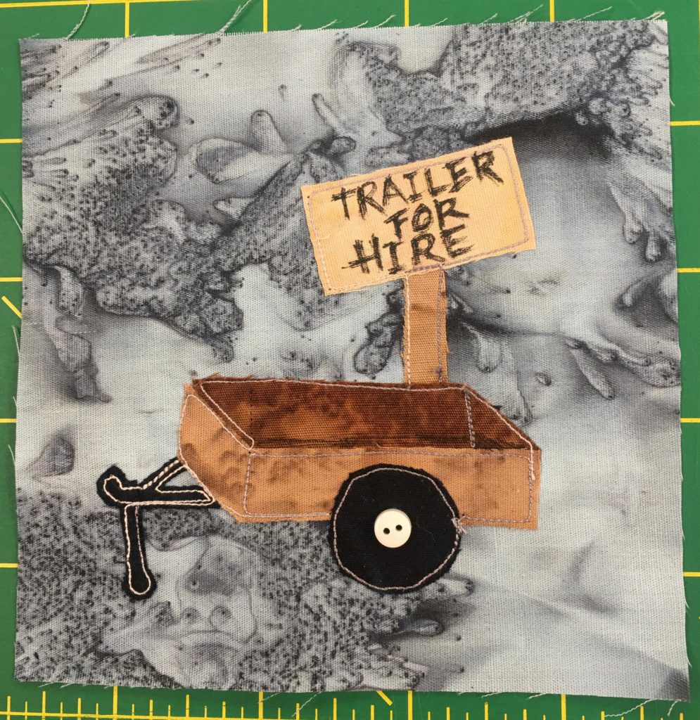 This quilt block shows a little box trailer that you might attach to a small car or truck against a gray background. The trailer is brown with a single wheel axis. The wheel is black with a small white button in the center to indicate it's attachment to the axis. There is a light brown wooden sign behind the trailer that looks like it's been written on by hand that reads,