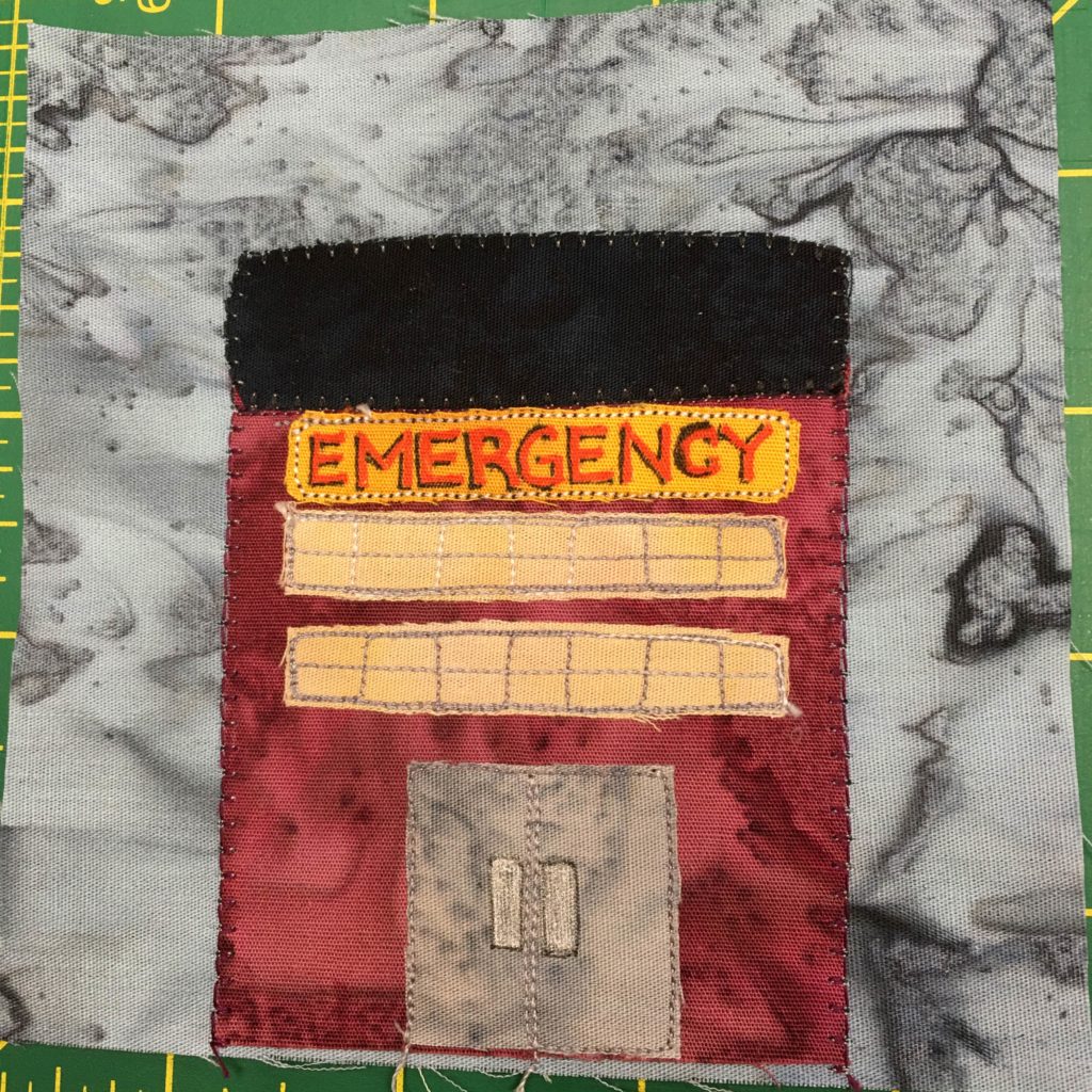 This quilt block shows a rectangular building on a gray background. The building has a black top, and a red exterior, like brick. There is a yellow sign across the top with red letters spelling out,
