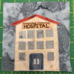 This quilt block shows a tall hospital building with a thing red sloped roof. The building is a little over three stories tall, and on the front is a grid of gray windows. At the bottom in the center is a pair of double-doors much like the Emergency Room center yesterday. These are darker colored with large gray handles.