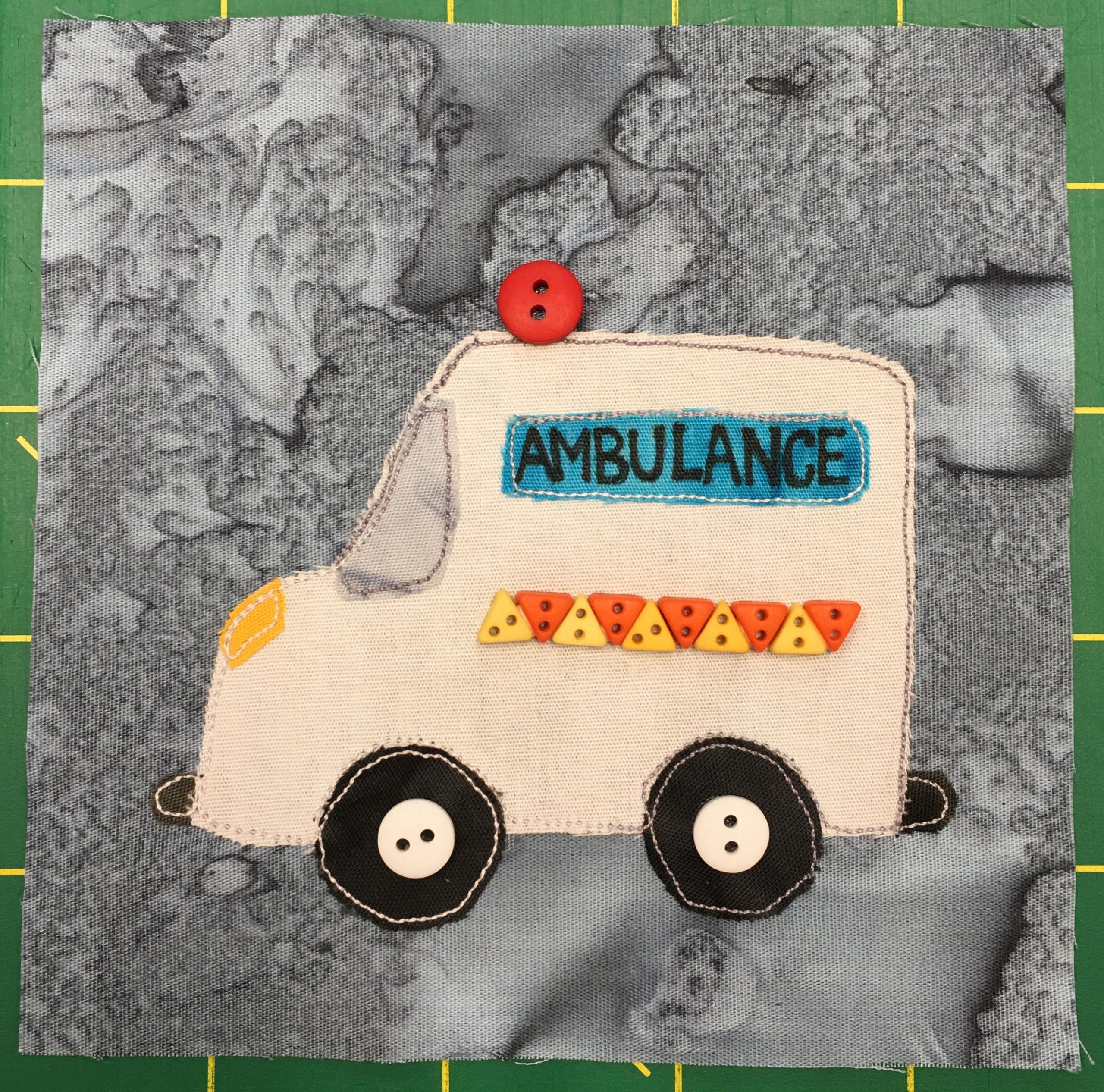 This quilt block shows am ambulance from the side with the driver cab facing to the left. It is white, with triangle buttons on it to indicate the shiny stripe on the carriage. There is a blue sign near the roof that reads, 