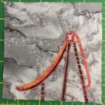 This quilt block shows an orange slide, with the same polkoa-dotted splotchy orange fabric that was used in previous park blocks. The chassis for the slide is a dark orange, while the slide and handles are brighter orange. The slide is a depicted sideways, so it's whole structure lies flat as if viewing it from the side with the end of the slide on the left.
