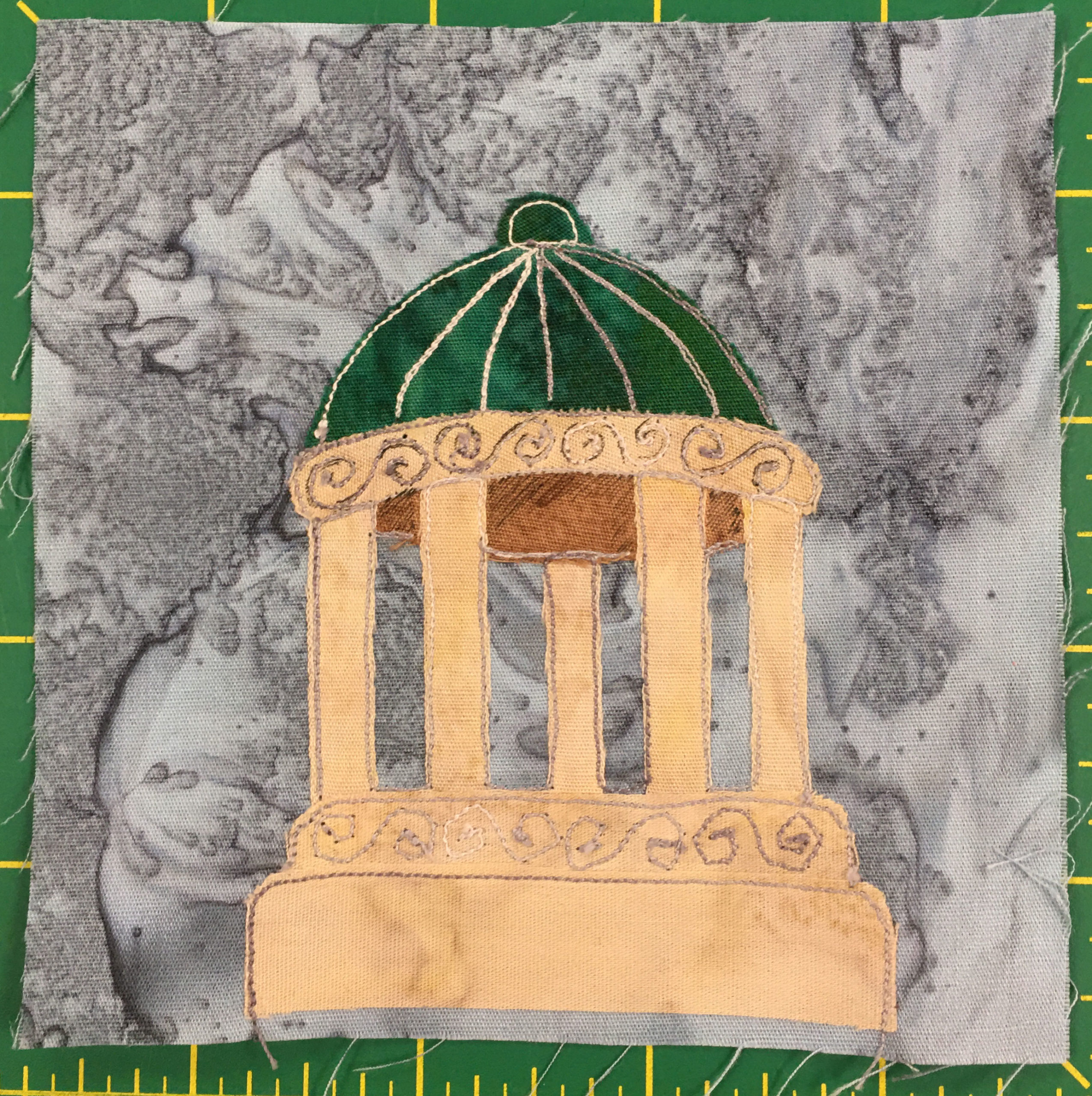 This quilt block shows a round gazebo or rotunda like structure with a base, columns, and a green dome top. The green dome top has white stitching on it cutting it into sections and a ball at the top of the dome. The columns are all in a circle supporting the dome, and below and above the columns is additional stone with decoration or etchings into the stone. The stone itself is a light yellow. The whole thing is very classically Greek like.