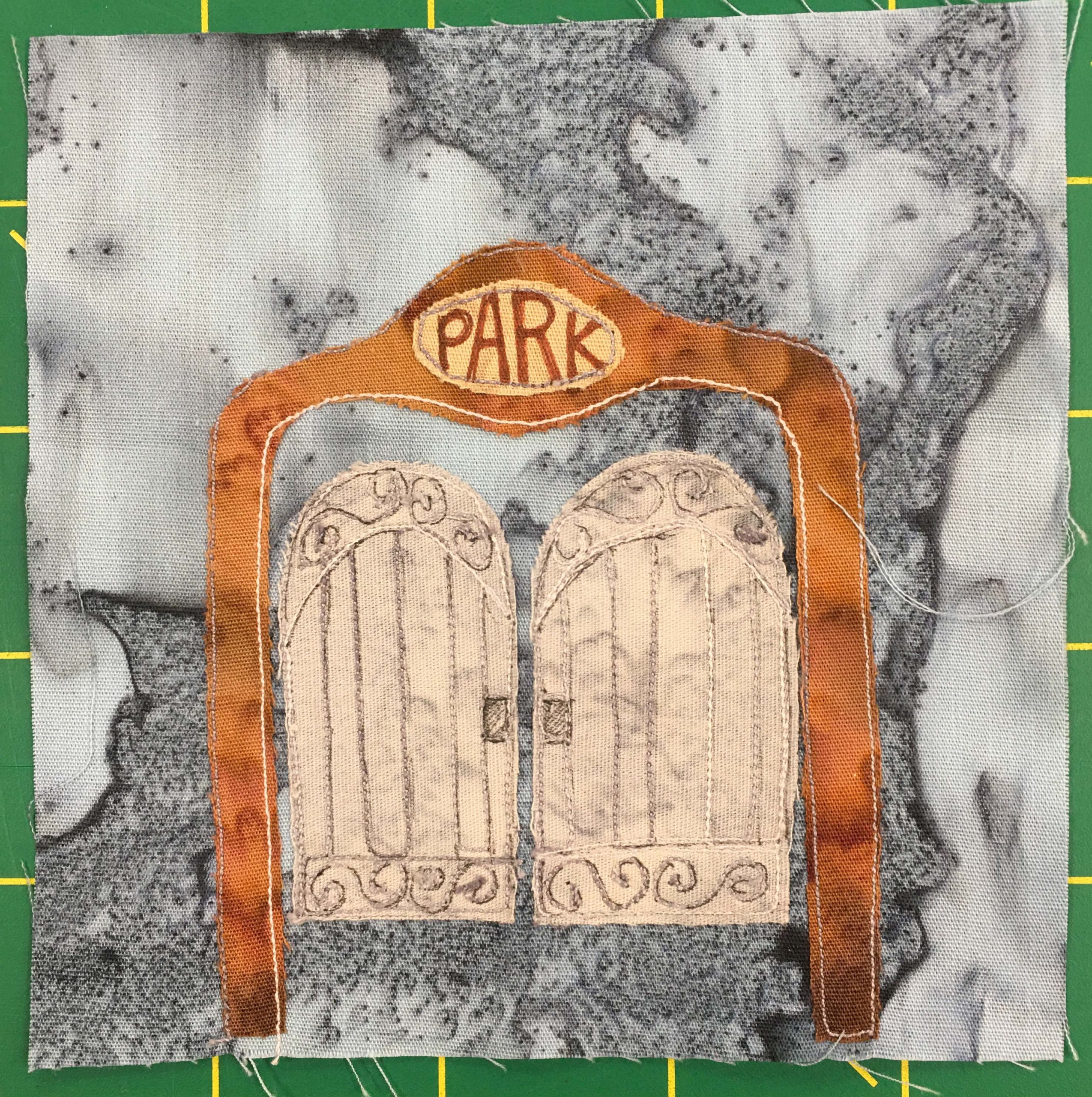 This quilt block shows a large gate against a gray background. The gate is huge, with a brown presumably metal frame forming an arc over the swinging double doors. Overhead, as you would walk under, is a sign in yellow that has the word, 