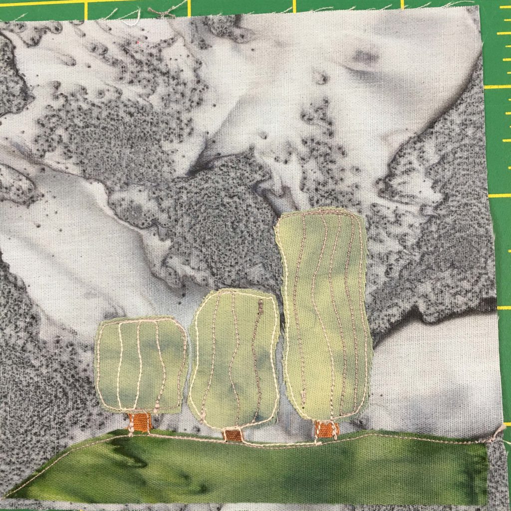 This quilt block shows a patch of smooth grass along the bottom with a deep green fabric, and then trees that look like light green blocks coming up from it. There are three trees with very short orange-brown trunks ranging from short to pretty tall. This is all against a gray salmon or fish like background fabric. There is vertical white stitching in the trees.