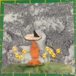 This quilt block shows a smooth patch of grass at the bottom denoted by a green cut of fabric. On the green fabric are threads shooting up with yellow buttons at their end indicating flowers. The buttons are round and two-holed. In the middle of it all is a rust orange light brown sundial stand, upon which sits a sundial. The stone bottom is segmented with thread, and a dark triangle, presumably in the shadow, stands erect in the center.