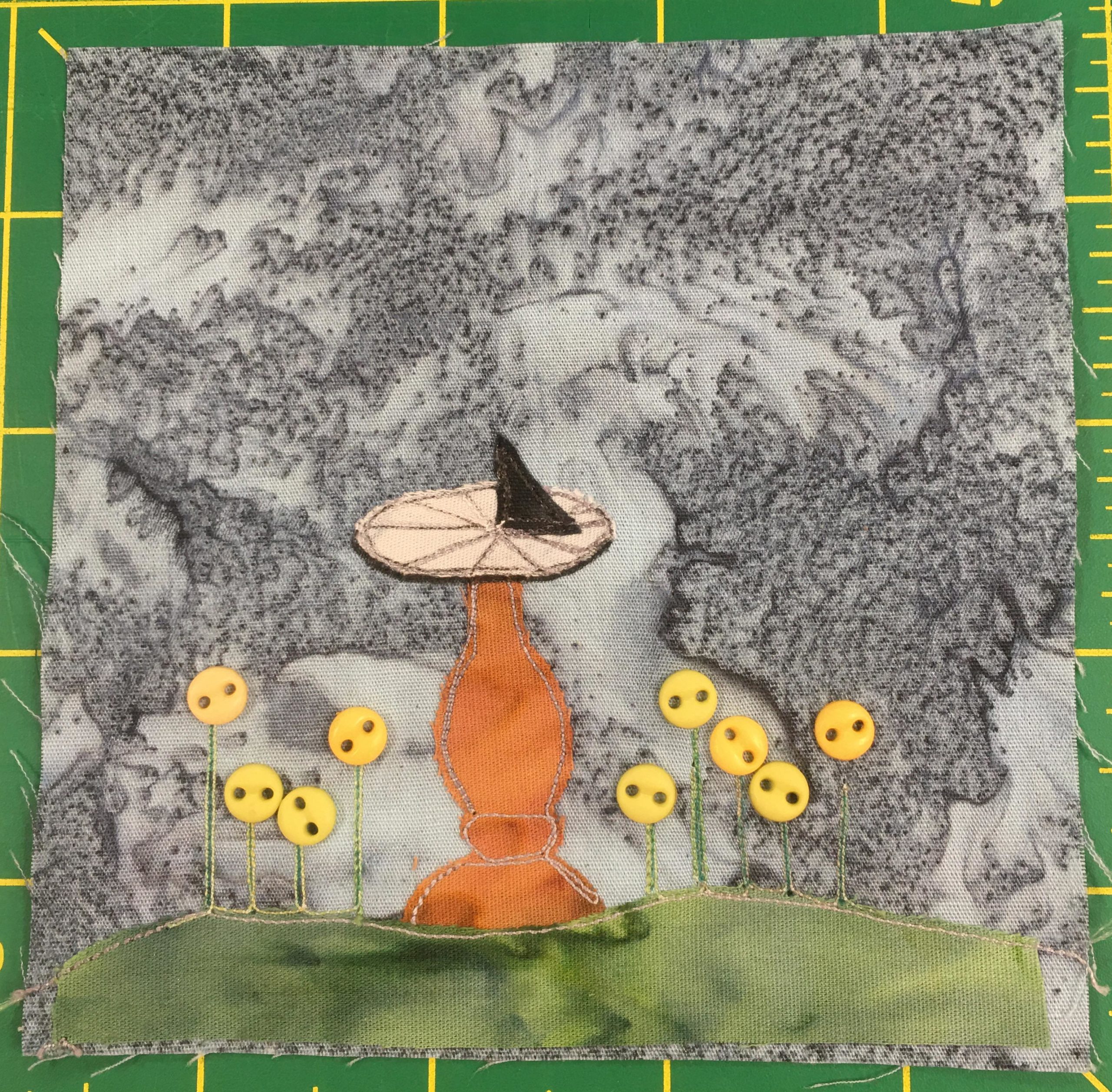 This quilt block shows a smooth patch of grass at the bottom denoted by a green cut of fabric. On the green fabric are threads shooting up with yellow buttons at their end indicating flowers. The buttons are round and two-holed. In the middle of it all is a rust orange light brown sundial stand, upon which sits a sundial. The stone bottom is segmented with thread, and a dark triangle, presumably in the shadow, stands erect in the center.