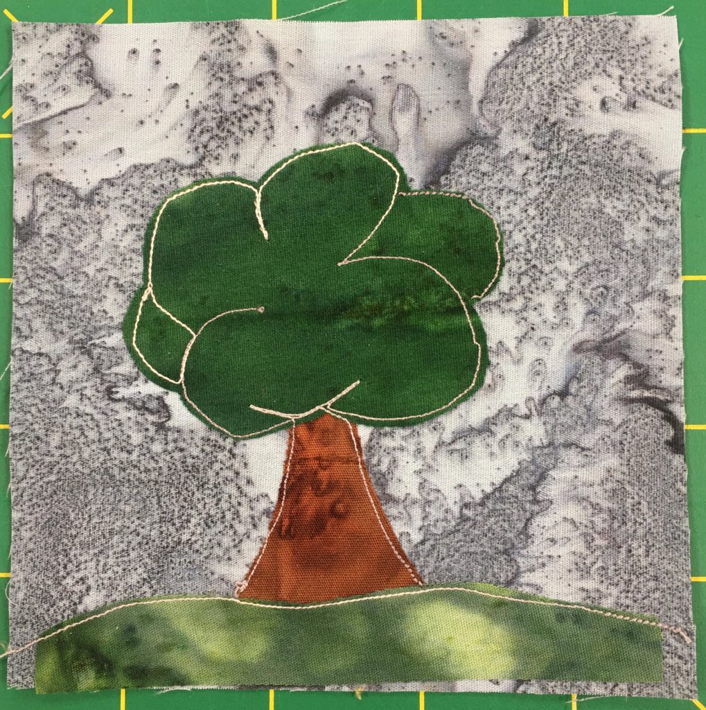 This quilt block shows a large pretty standard tree against a gray background. The tree is very large with billowy cloud like greenery at the top and a fat big trunk below. The trunk is a brown fabric, the grass a green splotchy fabric, and the greenery at the top a dark green with stitches to indicate cloud like bulges.