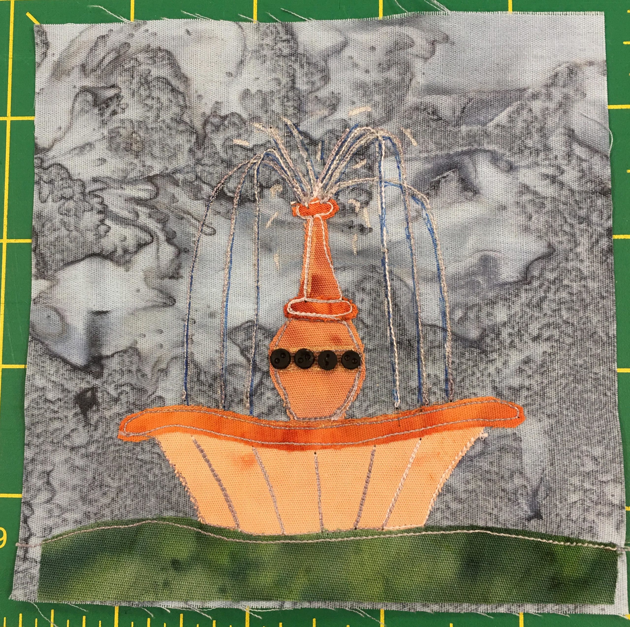 This quilt block shows a light orange fountain on a dark green patch of grass against a splotchy gray background. The fountain spout in the center has a bulb at the bottom with little miniature black buttons on it, and then narrows near the top where streams of water are shooting out (stitches and blue marker). The water lands in the shell like bottom pool.