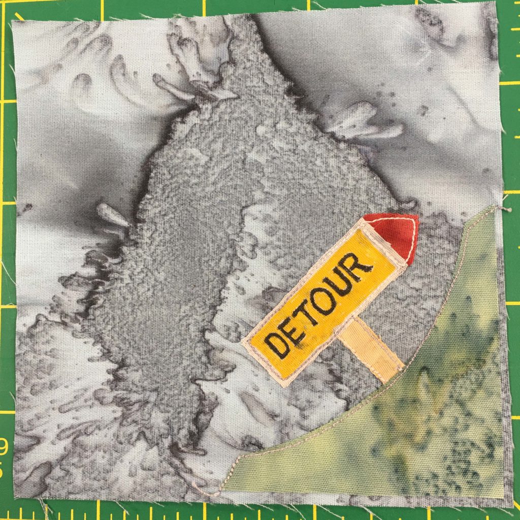 This quilt block shows a corner block. There is a patch of grass in the bottom right corner which doesn't extend to either the left or the top. At a forty-five degree angle on the patch of grass is a sign reading,
