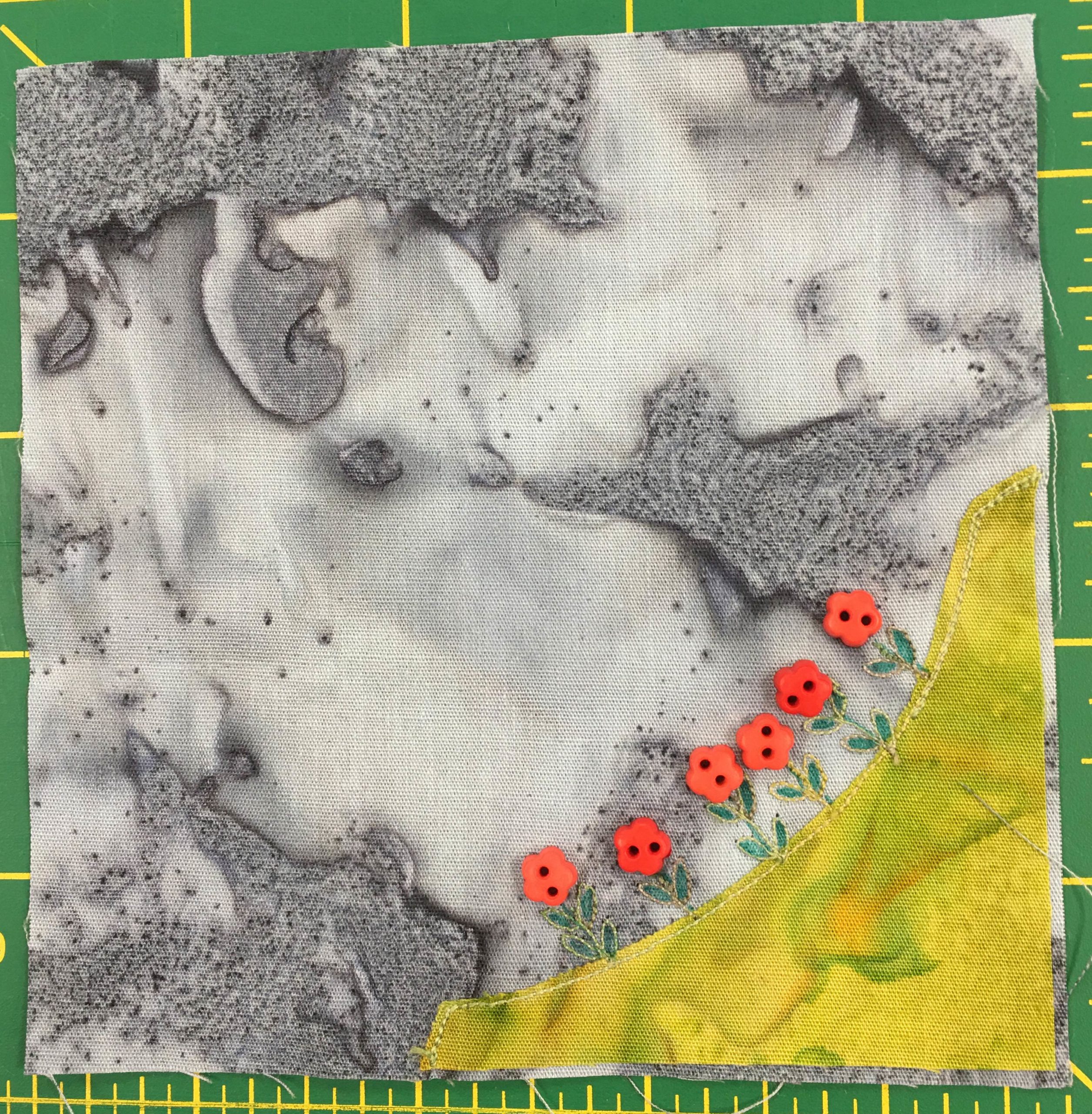 This is another corner quilt block. Against a gray background is a patch of grass in the bottom right, lighter than some of the other blocks before. There are little green sprouts, and on the sprouts are red two-holed flower shaped buttons.