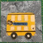This quilt block shows a yellow double-decker bus against a gray background. The bus has two big brown wheels with off-white two-holed round buttons in the centers. There is a driver's wheel penned in the bottom front of the bus, but more importantly there is a blue sign on the side that says,