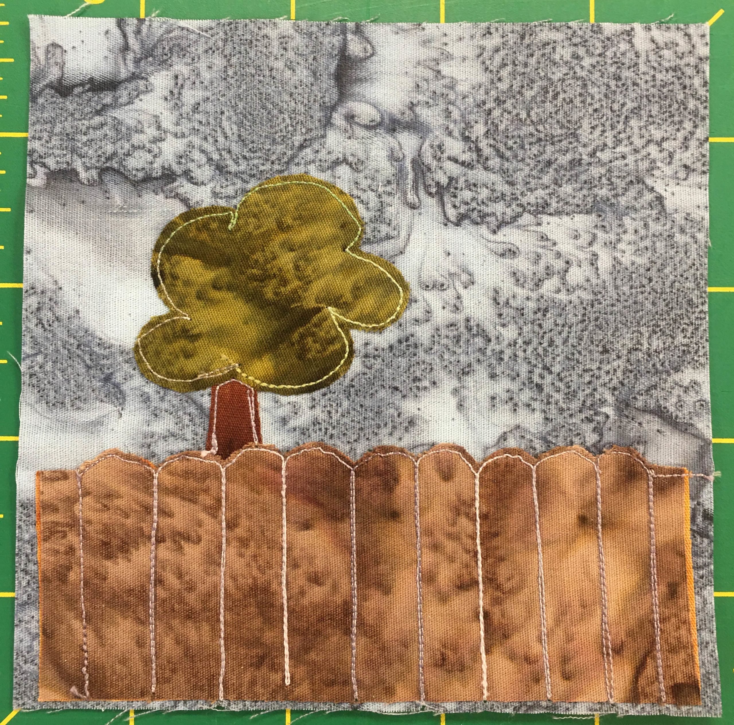 This quilt block shows a tree behind a fence against a gray background. The fence is a dark splotchy brown, and the top of each plank is rounded like a thumbnail. The fence takes up about one third of the block from the bottom, with the next third taken up by a dark rust-brown trunk and a somewhat blobby patch of dark green indicating a large tree.