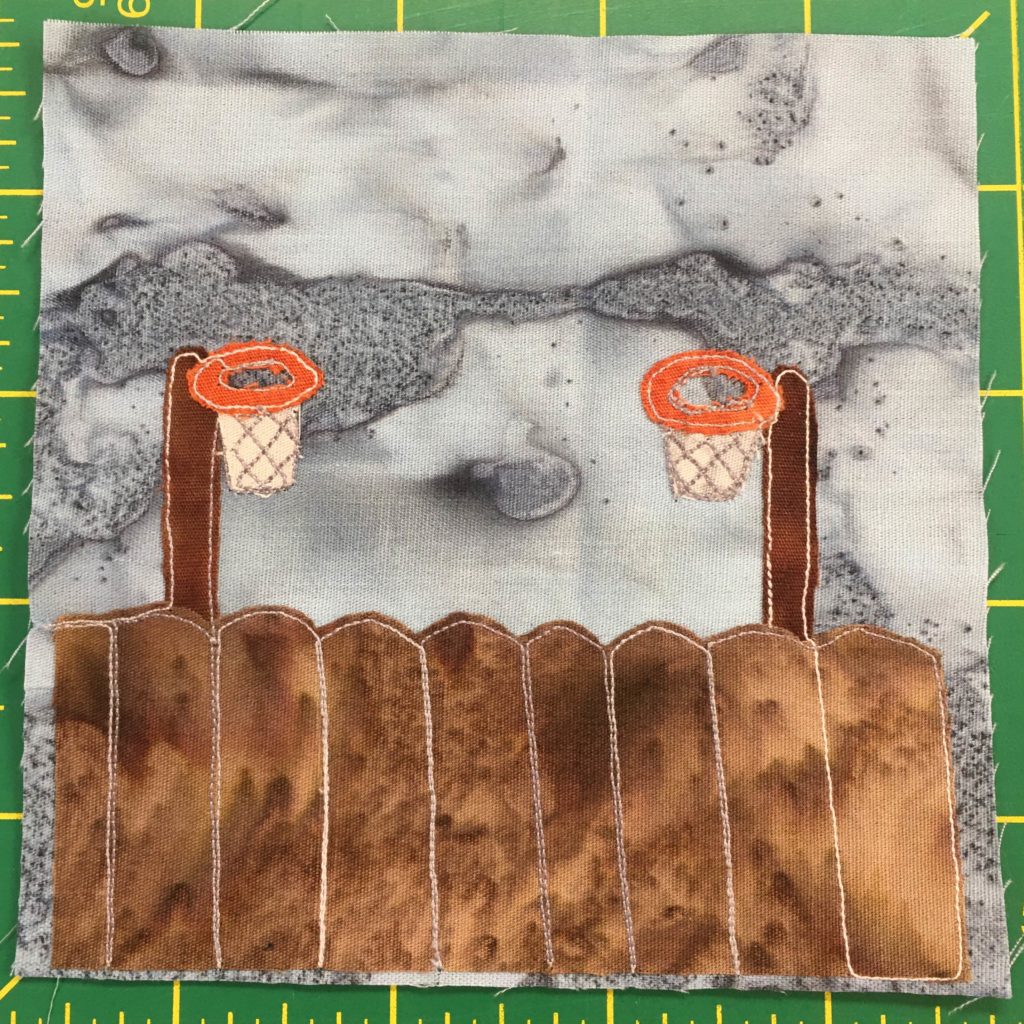 This quilt block shows a similar fence to the one in the previous block. Behind the brown rounded fence are two dark rust brown poles on either side of the block.  Attached to those two poles are bright orange circles with white fabric and thread under them indicating basketball hoops. This is all set against a splotchy gray background.