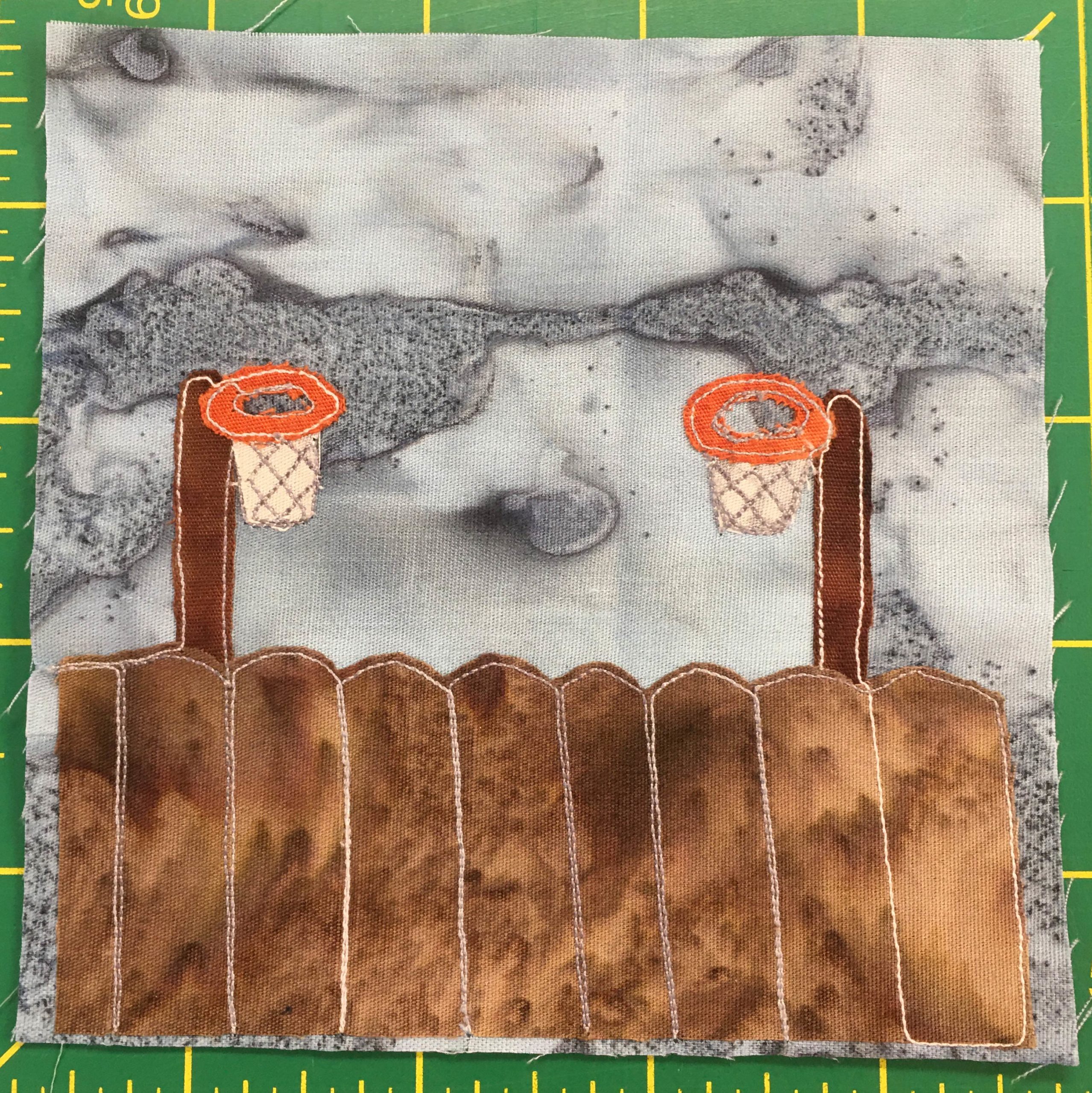 This quilt block shows a similar fence to the one in the previous block. Behind the brown rounded fence are two dark rust brown poles on either side of the block. Attached to those two poles are bright orange circles with white fabric and thread under them indicating basketball hoops. This is all set against a splotchy gray background.