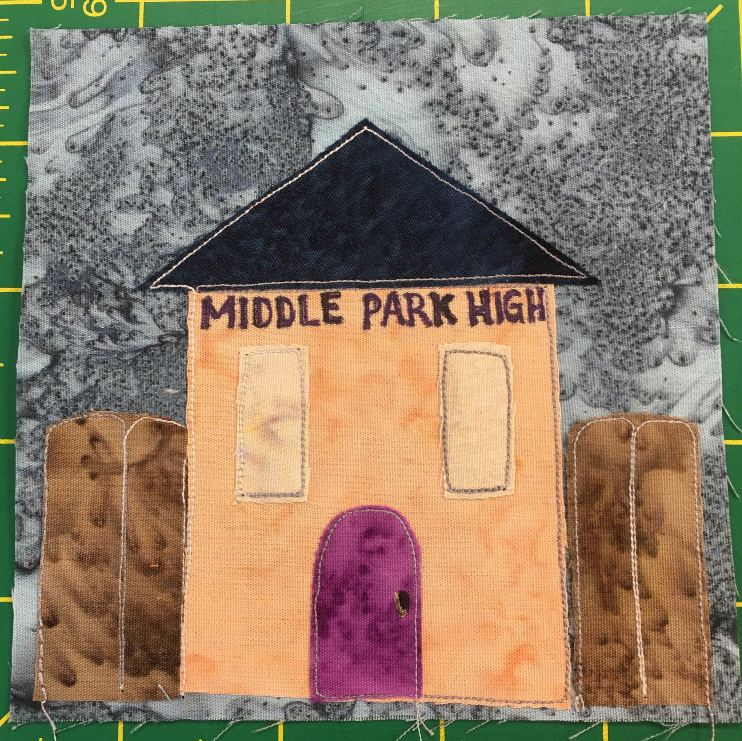 This quilt block has a light orange building in FRONT of the fence that appeared in the previous two blocks. The fence is a muddied brown with rounded edges that juts up about a third of the way of the block. The light orange building has a very dark triangle roof, and two long lighter orange windows on either side. There is a deep maroon/purple curved door below in the center. On the front of the building right under the roof are the words, 