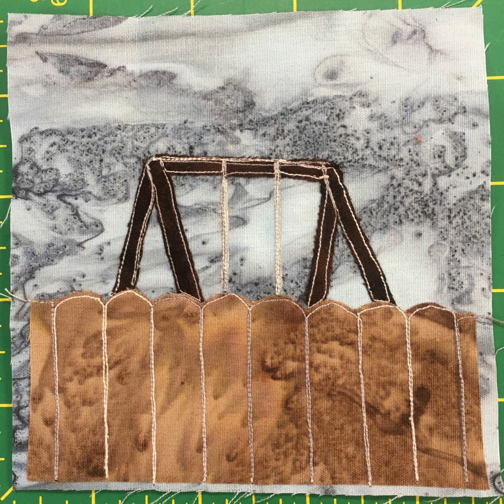 This quilt block shows a brown fence, like in the previous blocks, coming up just short of halfway. Above the fence you can see the top bars and chains of a swing-set. Both of these are against a gray splotchy background.