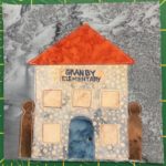 This quilt block shows a shortened fence, just two posts on either side of the center building. The center building is tall, two story, with a bright red triangular roof. On the front above the blue door and light yellow buildings is a sign that says, 