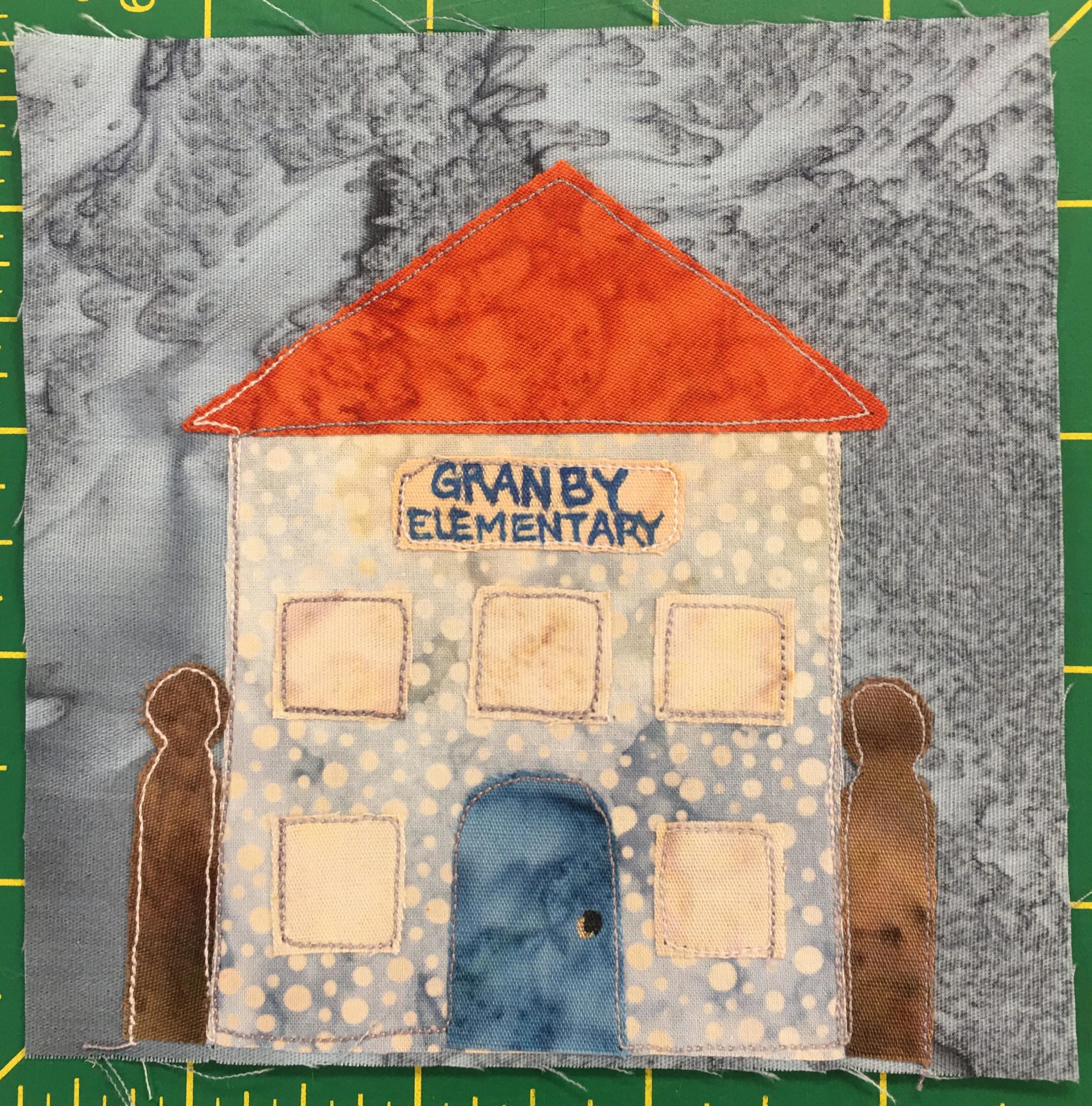 This quilt block shows a shortened fence, just two posts on either side of the center building. The center building is tall, two story, with a bright red triangular roof. On the front above the blue door and light yellow buildings is a sign that says, 
