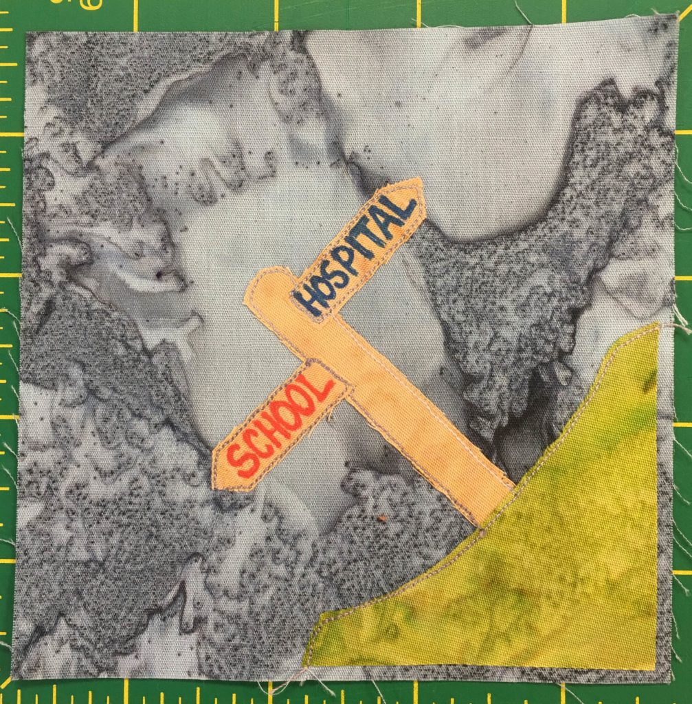 This quilt block shows a corner block, with a patch of bright green grass in the lower right corner. On the patch of grass is a light tan sign post with two signs sticking out in opposite directions. The signs are planks with pointed ends and read,