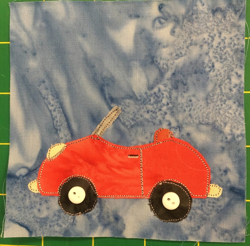 This quilt block shows a red little buggy convertible car with the roof off against a light blue background. The car itself is from a side view, with two dark wheels whose hubcaps are represented by two small white two-holed buttons. The red car only has one set of doors and no back seat.