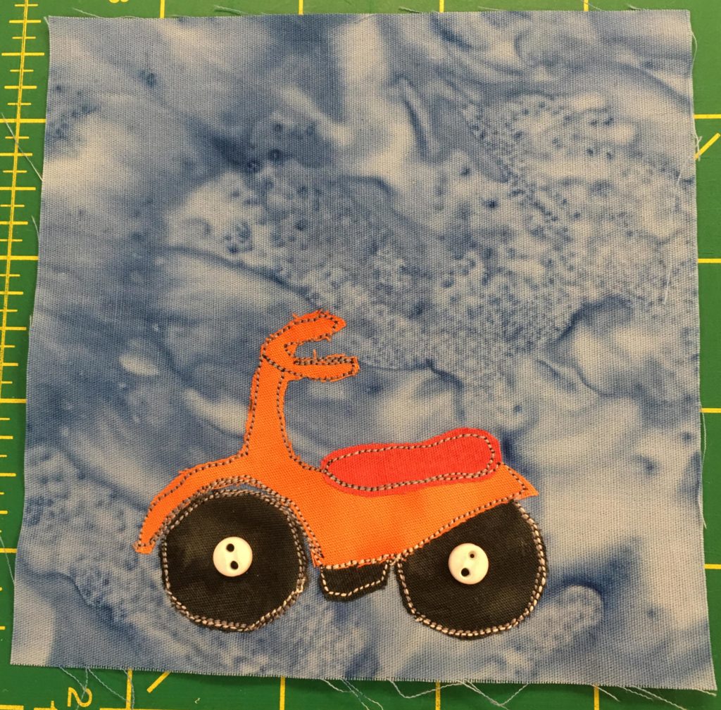 This quilt block shows an orange and red scooter like vehicle. It is shaped somewhat like a cross between a vespa and a motorcycle. This vehicle actually has three wheels however, with the bottom of the third wheel visible under the middle. The seat is red, and tehre are two handlebars. It is facing left, with two big dark tires whose axles are indicated by two white two-hole buttons.