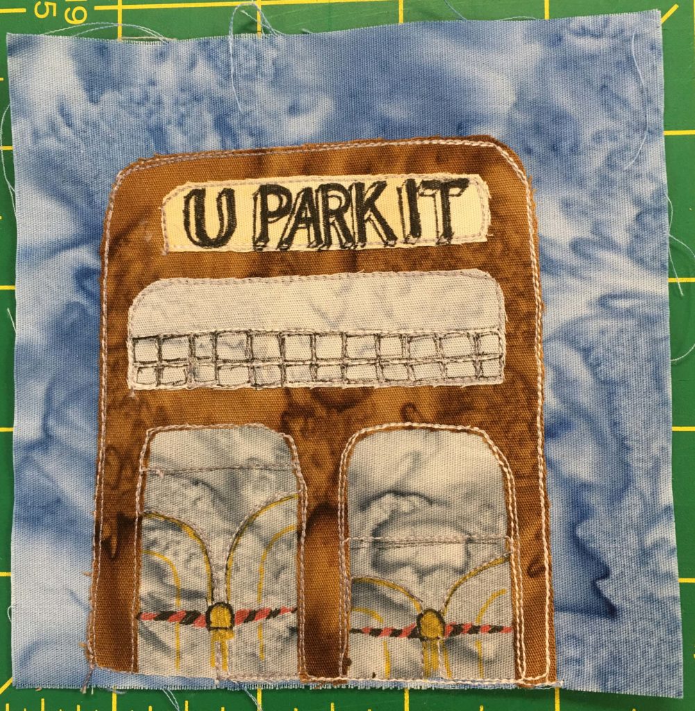 This quilt block shows a large brown building with a lower entrance level, and an upper level with a mesh metal railing. The yellow sign at the top against the muddy brown says,