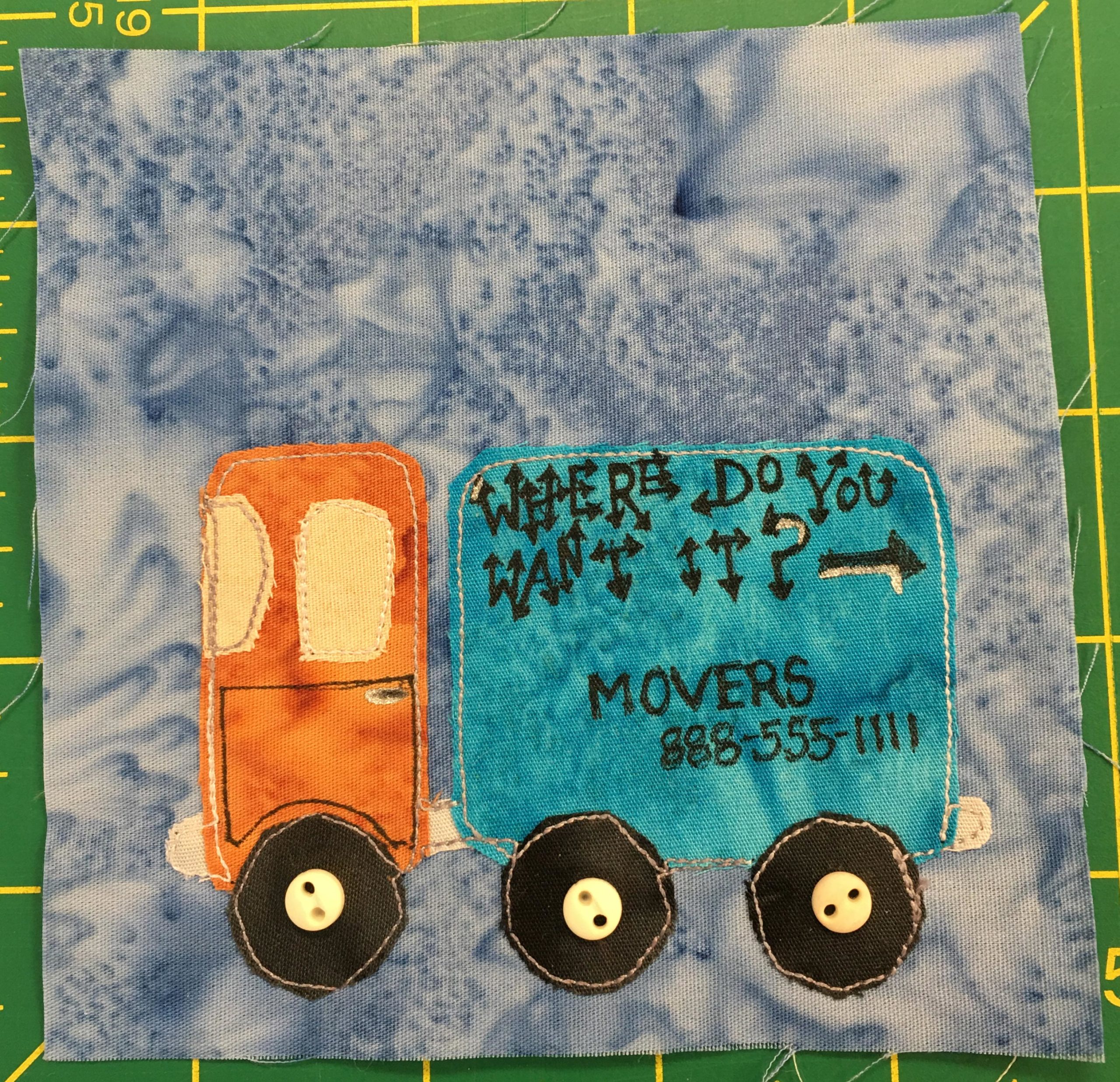 This quilt block shows a large two part truck. It's one truck, but it has a front cab, and a large back end for moving things. The front cab is bright orange, while the back end is a bright light blue. On the back end are the words, 