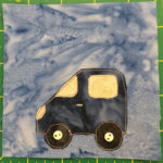 This quilt block shows a dark blue somewhat short compact car from a side view. It's facing left. The front angles down, and the back is a rectangle. The headlights, windshield, and window are yellow pieces of fabric. There are two tires that are black with off-white two-holed buttons in the center.