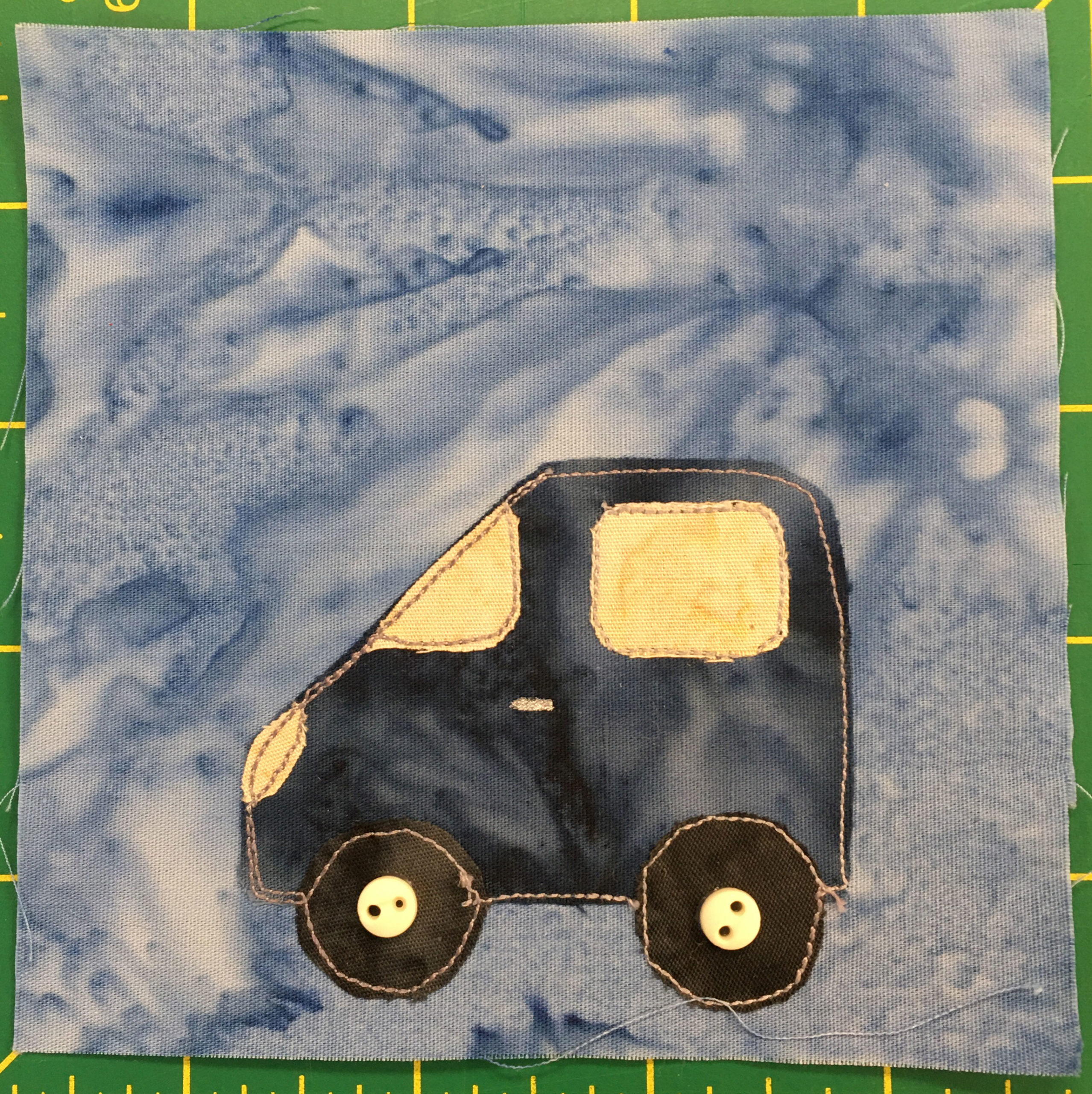This quilt block shows a dark blue somewhat short compact car from a side view. It's facing left. The front angles down, and the back is a rectangle. The headlights, windshield, and window are yellow pieces of fabric. There are two tires that are black with off-white two-holed buttons in the center.