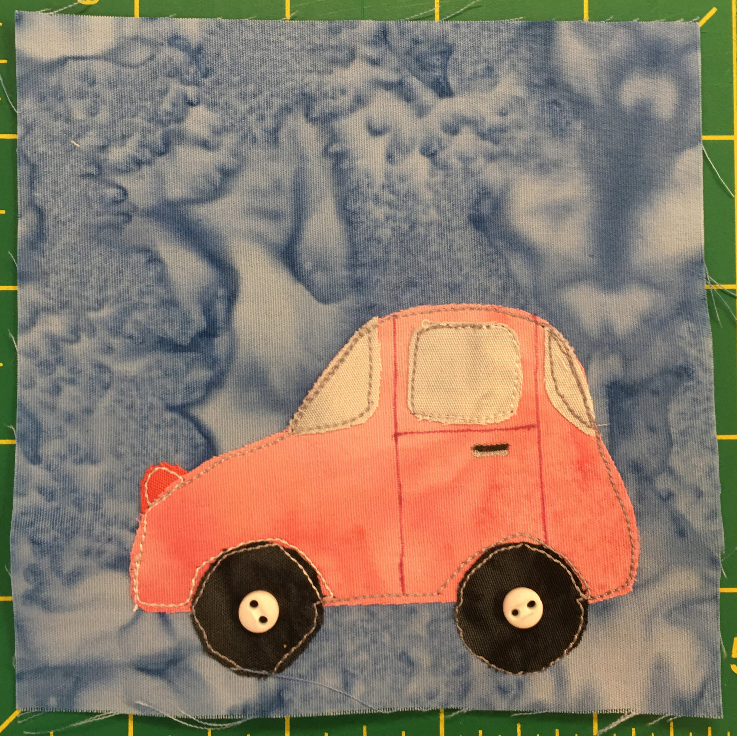 This quilt block shows another car but this time it is very pink. This car is rounder and you can see the rear window and a very small trunk. The headlight on the front of the car is a darker pink. The windows are a gray-yellow, and there are two dark wheels at the bottom. Each wheel has a small off-white two-hole button indicating the axle.
