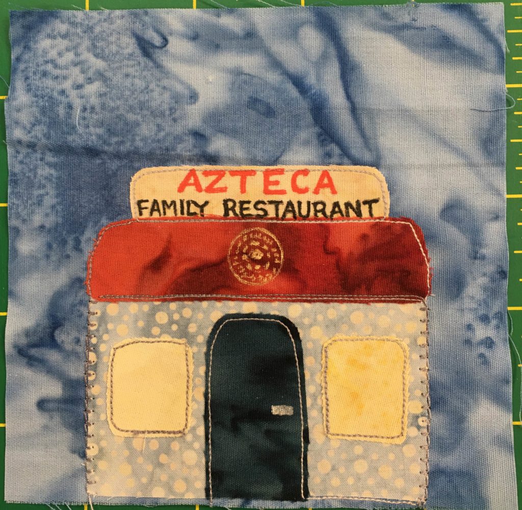 This quilt block shows a square building with a sign on top. The sign is light yellow and has the word