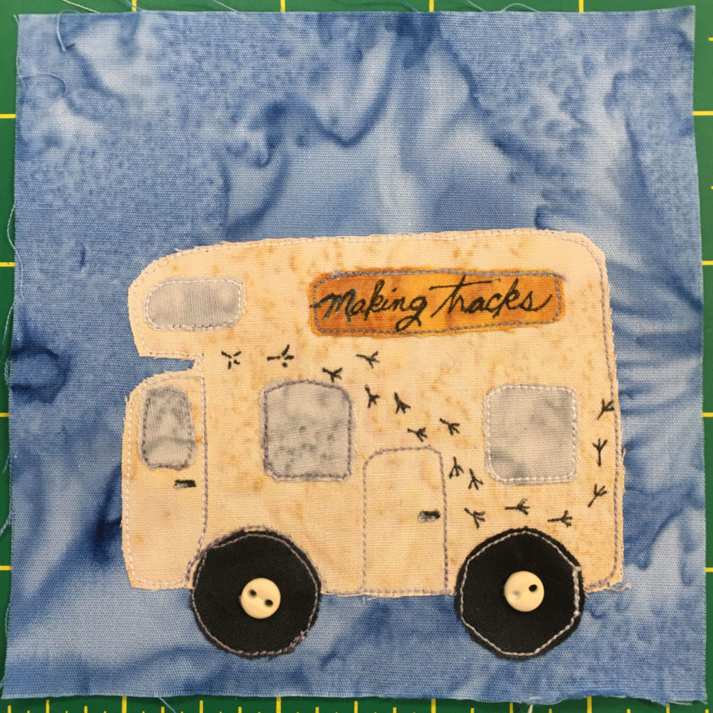 This quilt block shows a yellow camper truck. It is from the side view. On the side is a winding trail of bird footprints and a gold sign with the text,