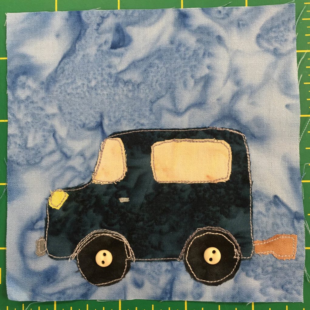 This quilt block shows a deep dark blue SUV against a light blue background (it's now day time in That Town). This vehicle is sturdy and has a tow hook on the rear of it (we are seeing it from the side.) The windshield and window are a light yellow and the headlight is a bright yellow. There are two dark wheels at the bottom with a small two-holed off-white in the center of each indicating the axle.