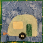 This quilt block shows a camper from the side view on a light blue background. The camper is a pale green with a dark green door on the right back side (behind the wheel). The window to the left is a light yellow. The camper is pill shaped a bit, with a rounded roof. There is a single wheel with a light two-holed small button in the middle of the dark circle indicating the axle.
