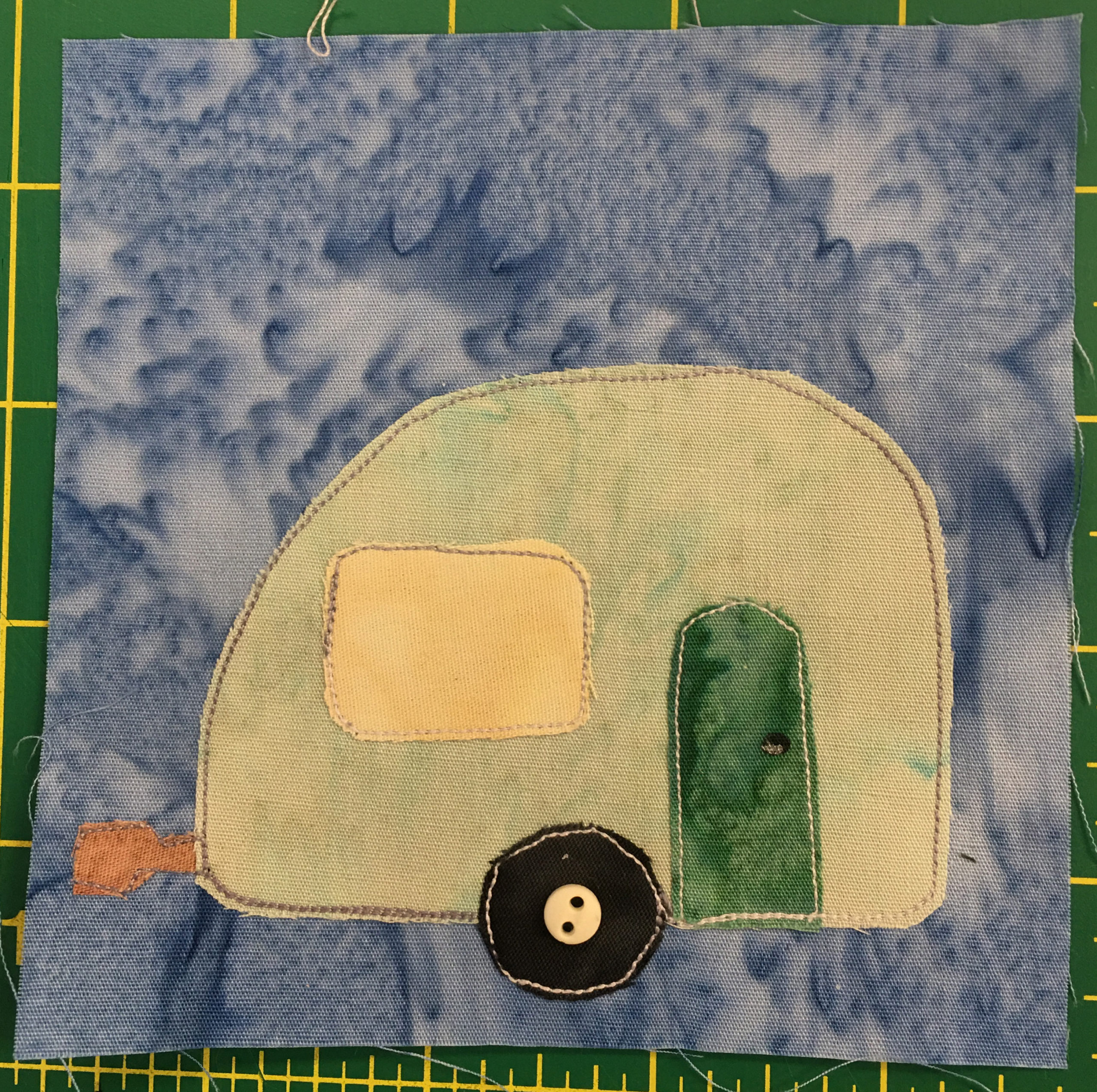 This quilt block shows a camper from the side view on a light blue background. The camper is a pale green with a dark green door on the right back side (behind the wheel). The window to the left is a light yellow. The camper is pill shaped a bit, with a rounded roof. There is a single wheel with a light two-holed small button in the middle of the dark circle indicating the axle.