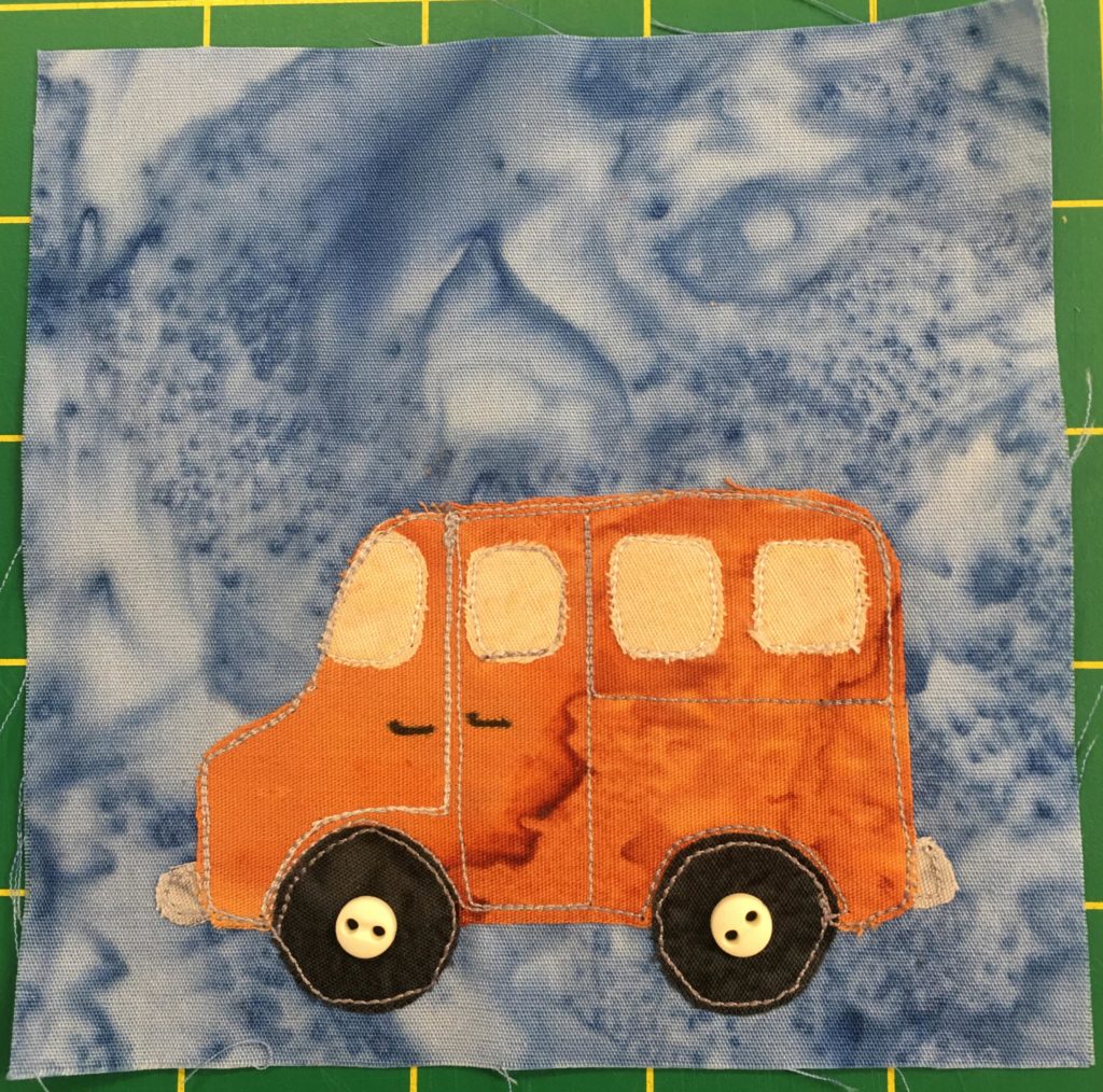 This quilt block shows a very orange van against a light blue background. The van has light yellow glass including windshield and windows. There are three windows on the side, one on a door and two as the carriage extends further back. There are two wheels below each with the obligatory small light two-holed buttons indicating the axles.
