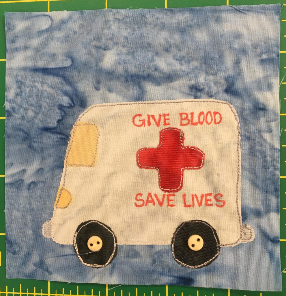 This quilt block shows a large ambulance like vehicle without lights against a light blue salmon-patterned background. The vehicle isn't an emergency vehicle, but is large and white and on the side has a big red plus sign. There are also the words above and below that read,