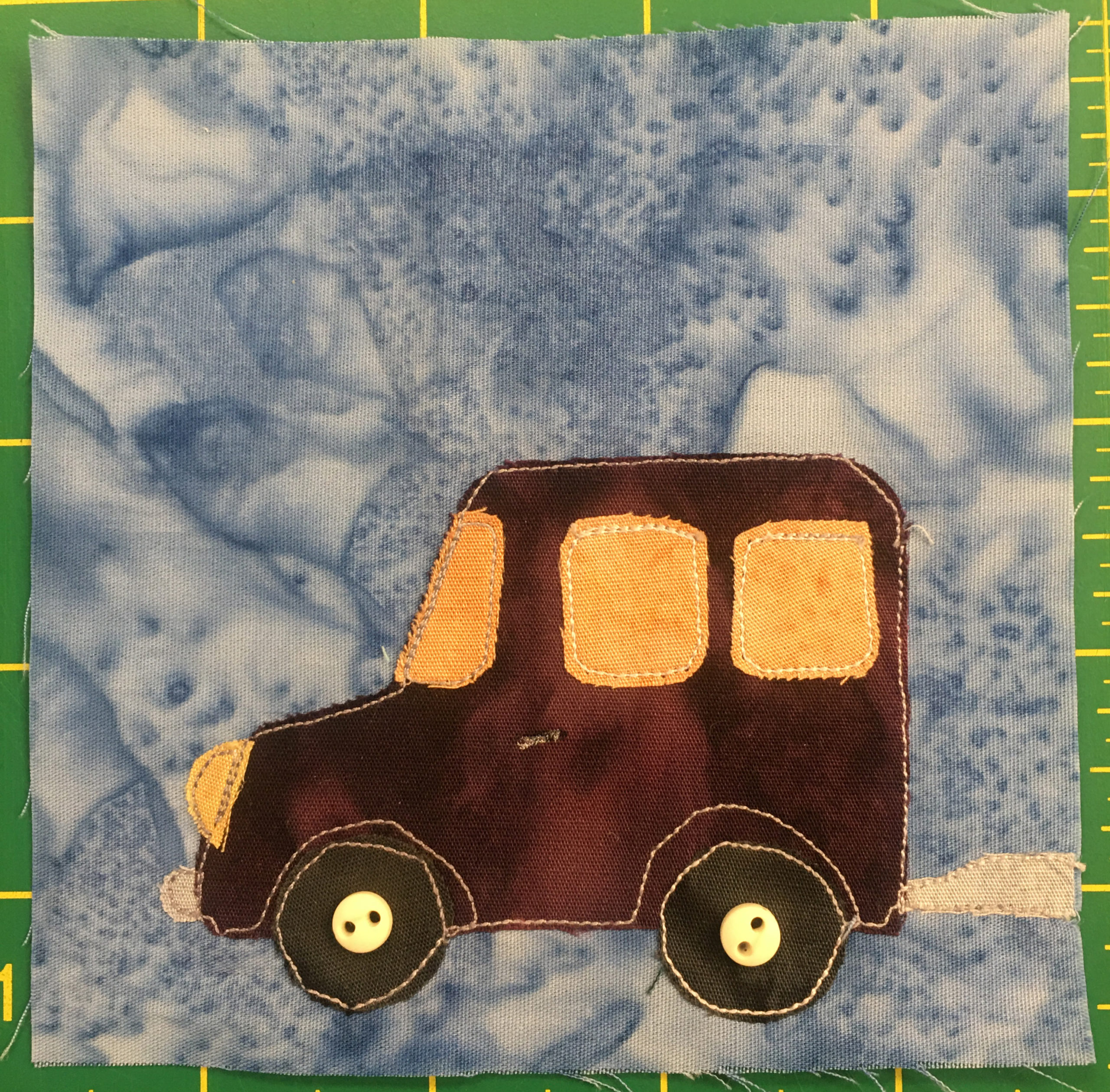 This quilt block shows a dark brown and slightly reddish truck without a bed. The truck's can extends to the rear of the truck and it has two windows light orange windows besides the windshield. The headlight is also the same color. Behind the truck is a gray tow structure attached to something that's located off the quilt block to the right (the truck is facing left.) There are two dark circles indicating wheels underneath with small off-white two-holed buttons in the centers indicating the axle.