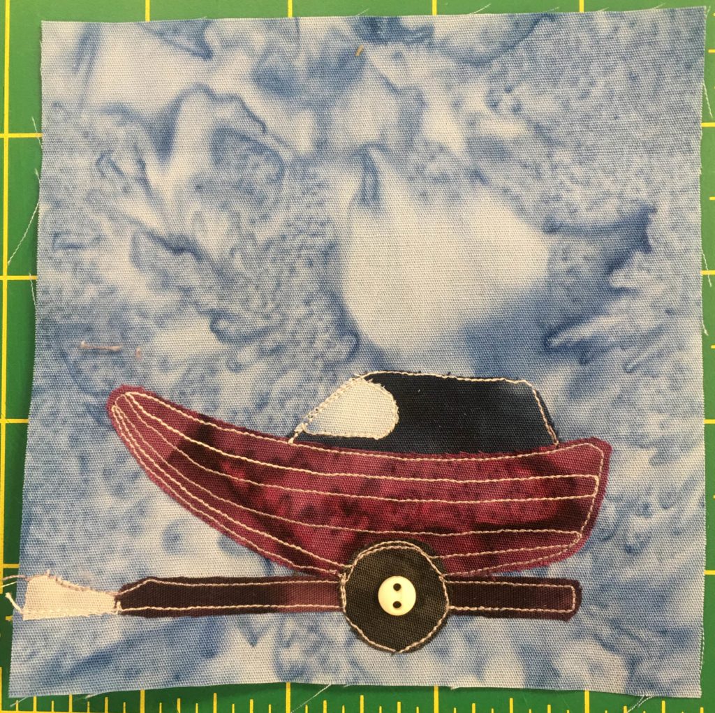 This quilt block shows a maroon boat on a trailer against a light blue salmon-patterned background. The boat hull has light thread lines stitched through it, and is made out of a dark maroon piece of fabric,. The top of the boat is dark blue with a windshield opening that is light blue. It's sitting on a dark brown trailer with a single wheel that is a dark circle with a small off-white two-holed button in the middle indicating the axle.