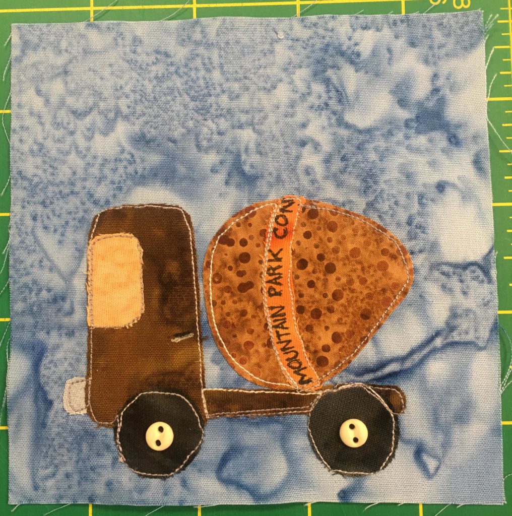 This quilt block shows a brown and orange cement truck against a light blue salmon-patterned background. The cab is brown with a light orange windshield. The fabrics are splotchy except for the mixer which is orange with brown speckled polka-dots. There is an orange band around the mixer with writing that says,