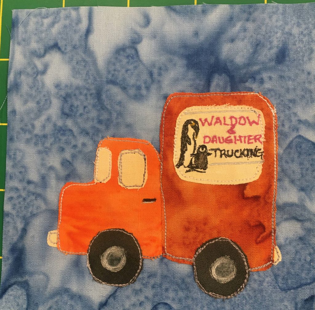This quilt block shows a bright orange truck on a light blue salmon-patterned background. The cab of the truck is a brighter orange than the bed of the truck, which is covered like a big rig. The wheels are dark circles with white hubcaps drawn on by pen. There is a big sign on the tall cab on the truck that shows a adult penguin taking care of a baby penguin. There are pink words that read, "Waldow & Daughter" and underneath it in black, "Trucking."