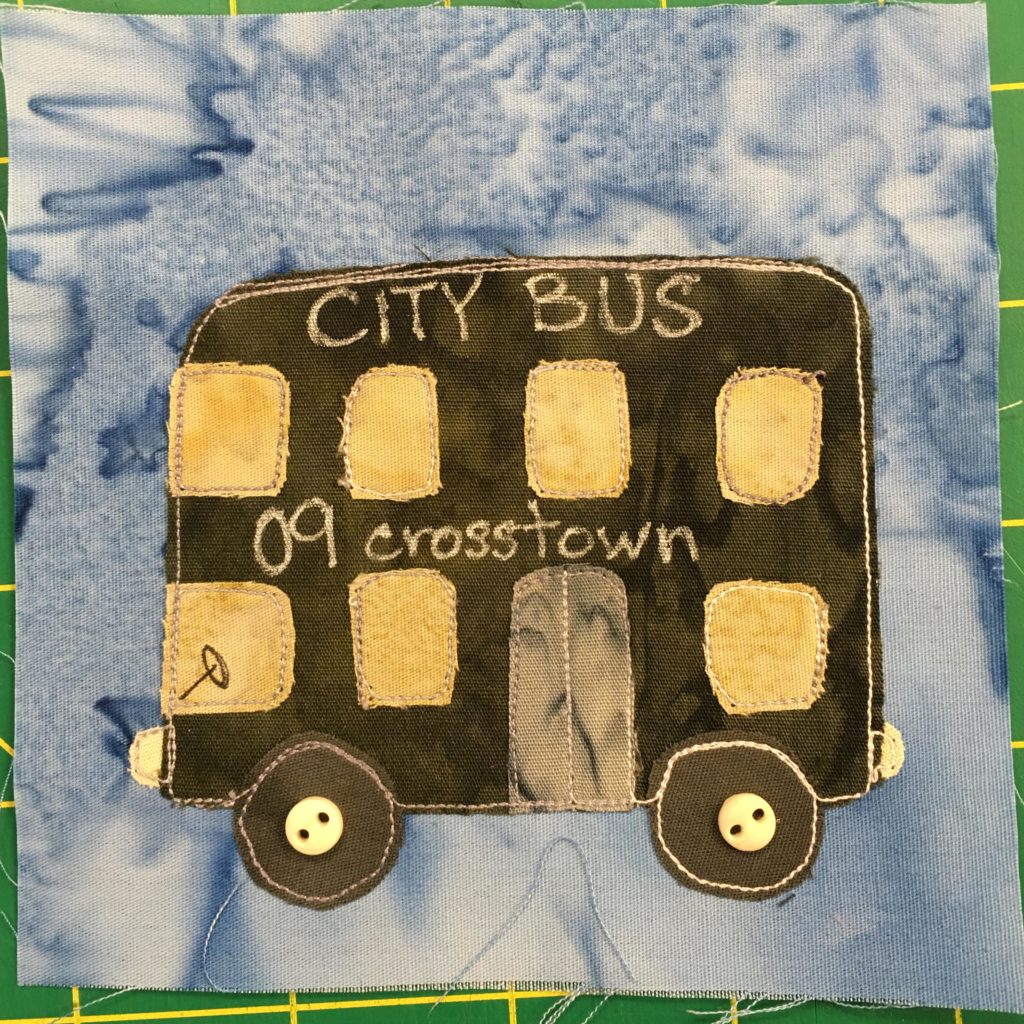This quilt block shows a large double decker bus that's a dark off-green. The windows are a light yellow. There are four across the top and three on the bottom, interrupted by the gray door that opens in the middle. A steering wheel is visible through the lower cab window. There are words penned in on the side of the bus,