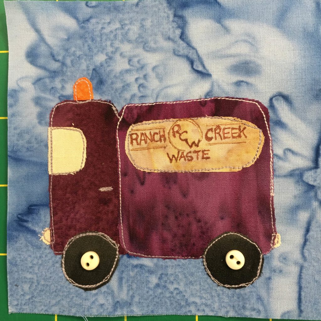 This quilt block shows a purple or deep marron square truck whose bed is as tall and big as its cab. There is an orange warning light on top of the cab. The window of the cab is a light yellow. The bed has a yellow sign on the side with a logo that reads,