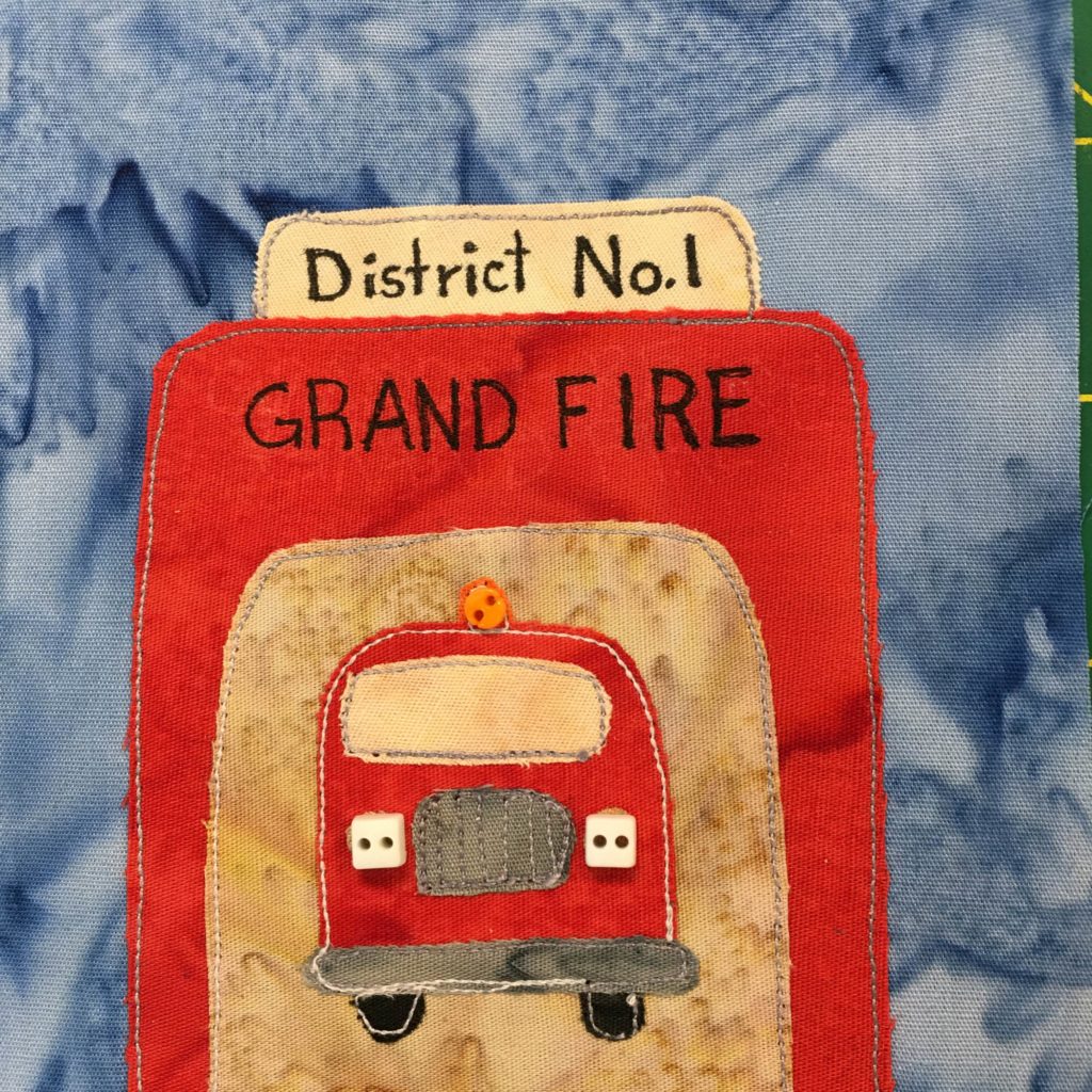 This quilt block shows a fire station garage with the door open against a deep blue salmon-patterned background. The station building itself is a bright red, the same shade as the fire engine which we can see from the front. The sign on top is yellow, and reads, 