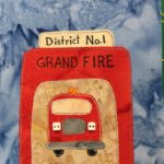 This quilt block shows a fire station garage with the door open against a deep blue salmon-patterned background. The station building itself is a bright red, the same shade as the fire engine which we can see from the front. The sign on top is yellow, and reads,